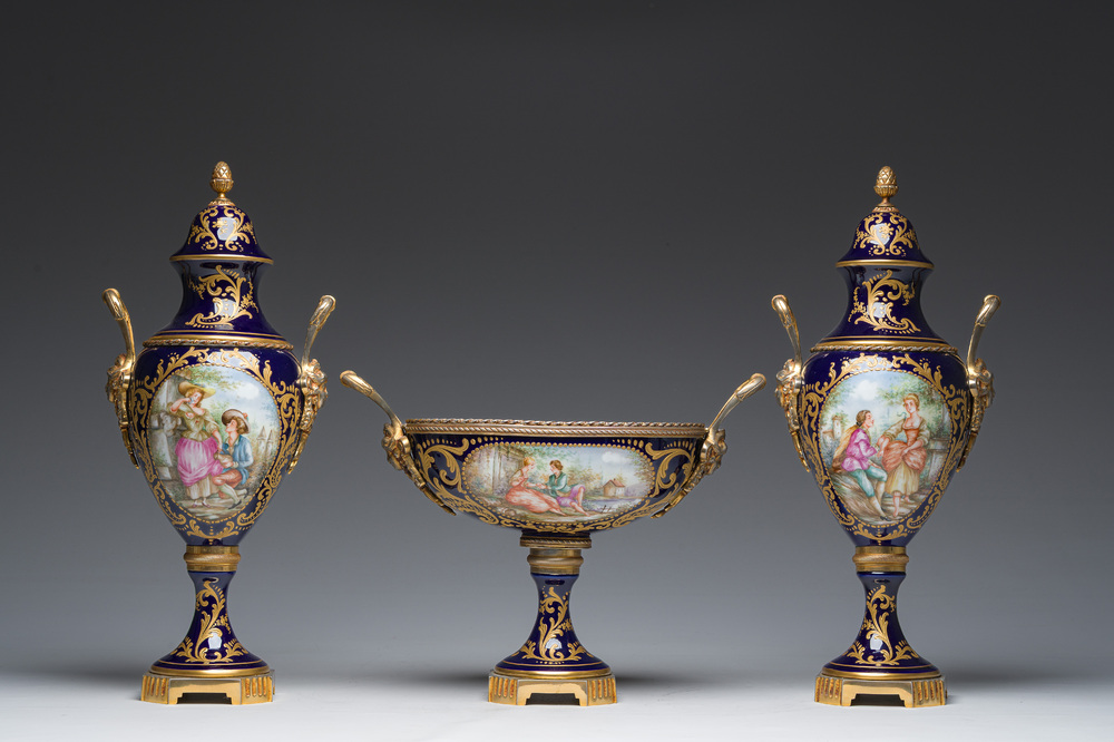 Een driedelige Franse S&egrave;vres-stijl schouwgarnituur met blauwe fondkleur, verguld bronzen monturen en galante sc&egrave;nes, 20ste eeuw