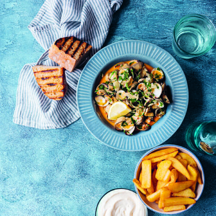 Bilden föreställer en blå tallrik på en blå bordskiva. I tallriken finns blåmusslor med nduja. Intill tallriken står en skål med pommes frites, en skål med aioli och rostat surdegsbröd.