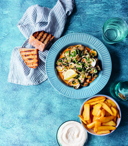 Bilden föreställer en blå tallrik på en blå bordskiva. I tallriken finns blåmusslor med nduja. Intill tallriken står en skål med pommes frites, en skål med aioli och rostat surdegsbröd.