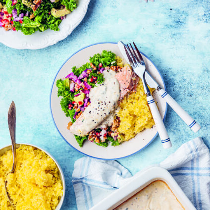 Bilden föreställer en vit porslinstallrik som står på en ljusblå bordsskiva. På tallriken ligger en laxfile täckt med en krämig vit sås med ädelost. Intill laxen finns pressad potatis och en färgsprakande i grönt och rött med granatäppelkärnor.