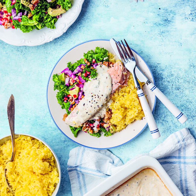 Bilden föreställer en vit porslinstallrik som står på en ljusblå bordsskiva. På tallriken ligger en laxfile täckt med en krämig vit sås med ädelost. Intill laxen finns pressad potatis och en färgsprakande i grönt och rött med granatäppelkärnor.